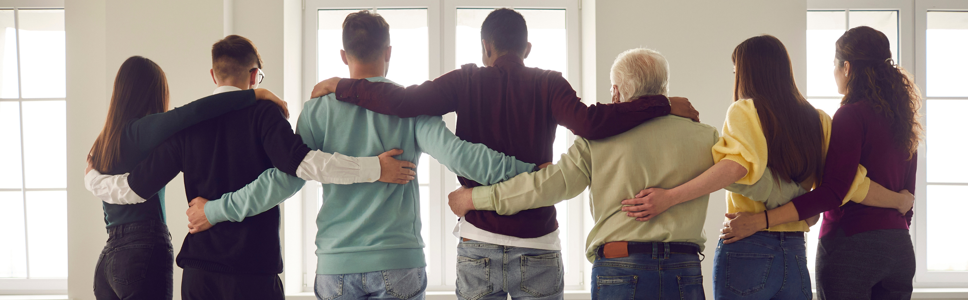 Team of People Hug Standing in a Row with Their Backs to the Camera and Looking Out the Window.